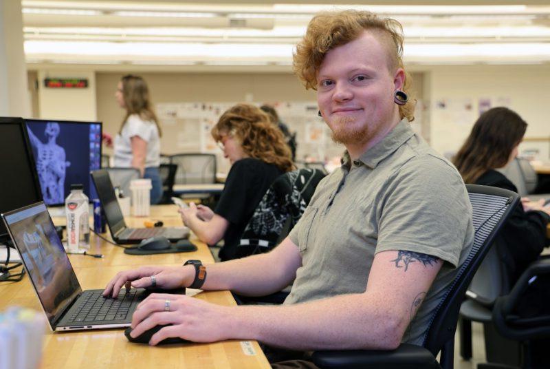 A student sits at a desk in front of an open laptop computer.